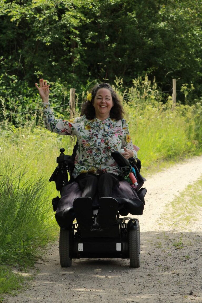 Riad foto af Tina der vinker i kørestol og blomstret bluse. Tilgængelighed med kørestol